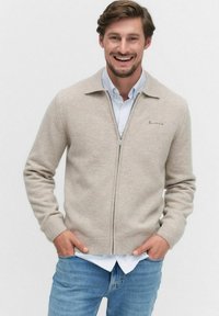 Beige zip-up kofta i mjukt material med krage, ribbade ärmslut och fåll. Lager på en ljusblå rutad skjorta.