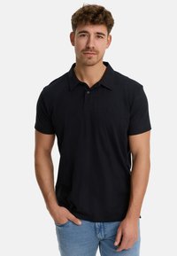 Camisa polo negra de algodón, con un cuello clásico, mangas cortas y un discreto detalle de logo en el lado izquierdo del pecho. Combinada con jeans de mezclilla azul claro.