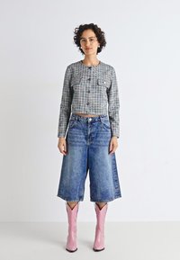Veste à carreaux grise, courte, avec fermeture à boutons, associée à un pantalon large en denim bleu et des bottes de cowboy roses brillantes.