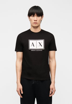 Tânăr cu păr negru, purtând un tricou negru Armani Exchange cu logo alb și pantaloni negri, stând în picioare pe un fundal simplu, deschis la culoare.