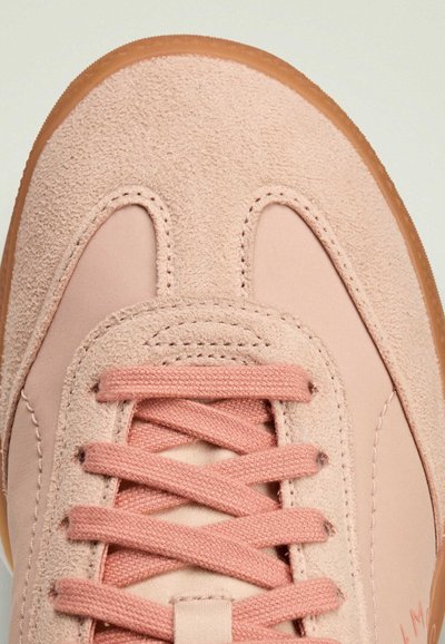 Light pink sneaker with a textured suede toe and smooth side panels. Features flat laces, a tan rubber sole, and subtle stitching details.