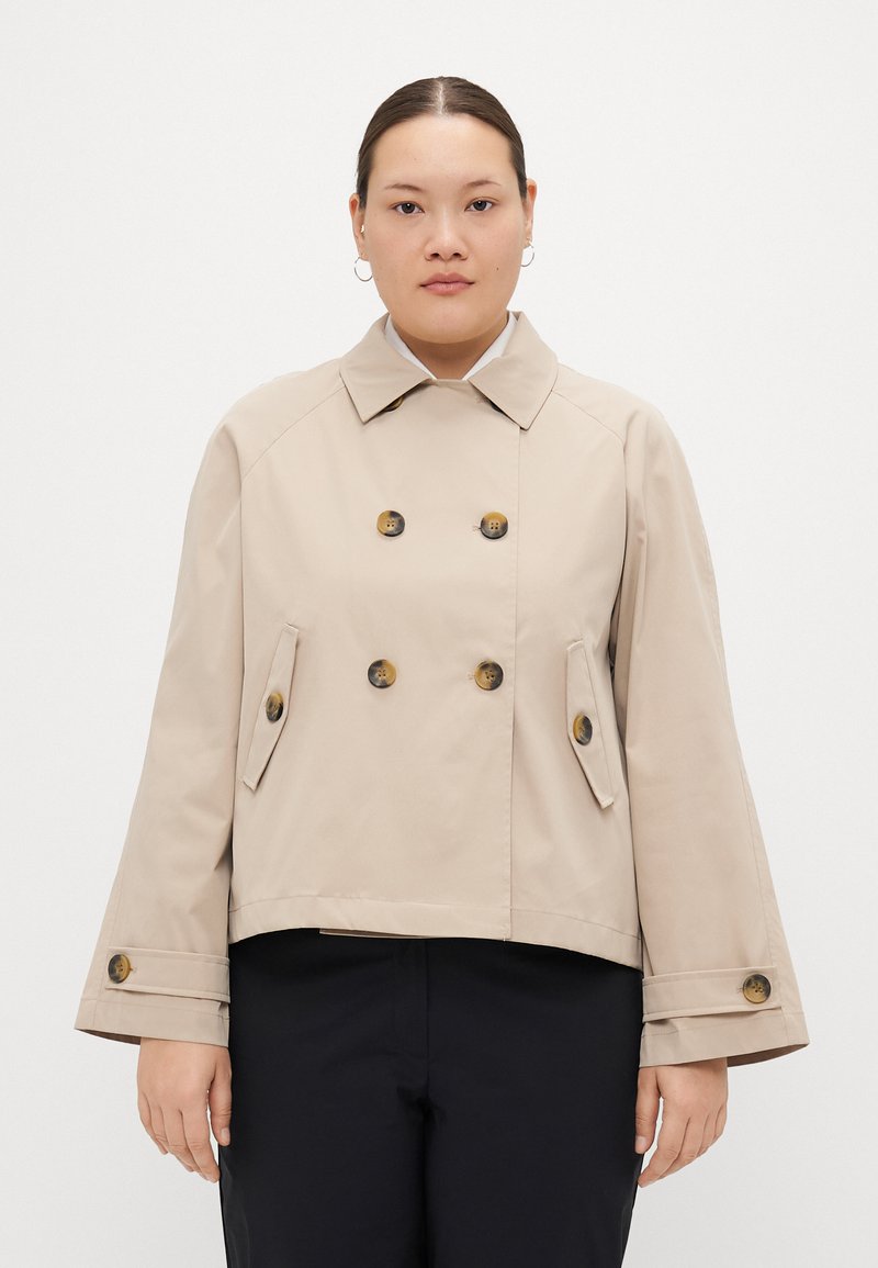 Beige kort jakke med dobbelspent knapper, skrå lommer og vide ermer. Laget av glatt, lett stoff.