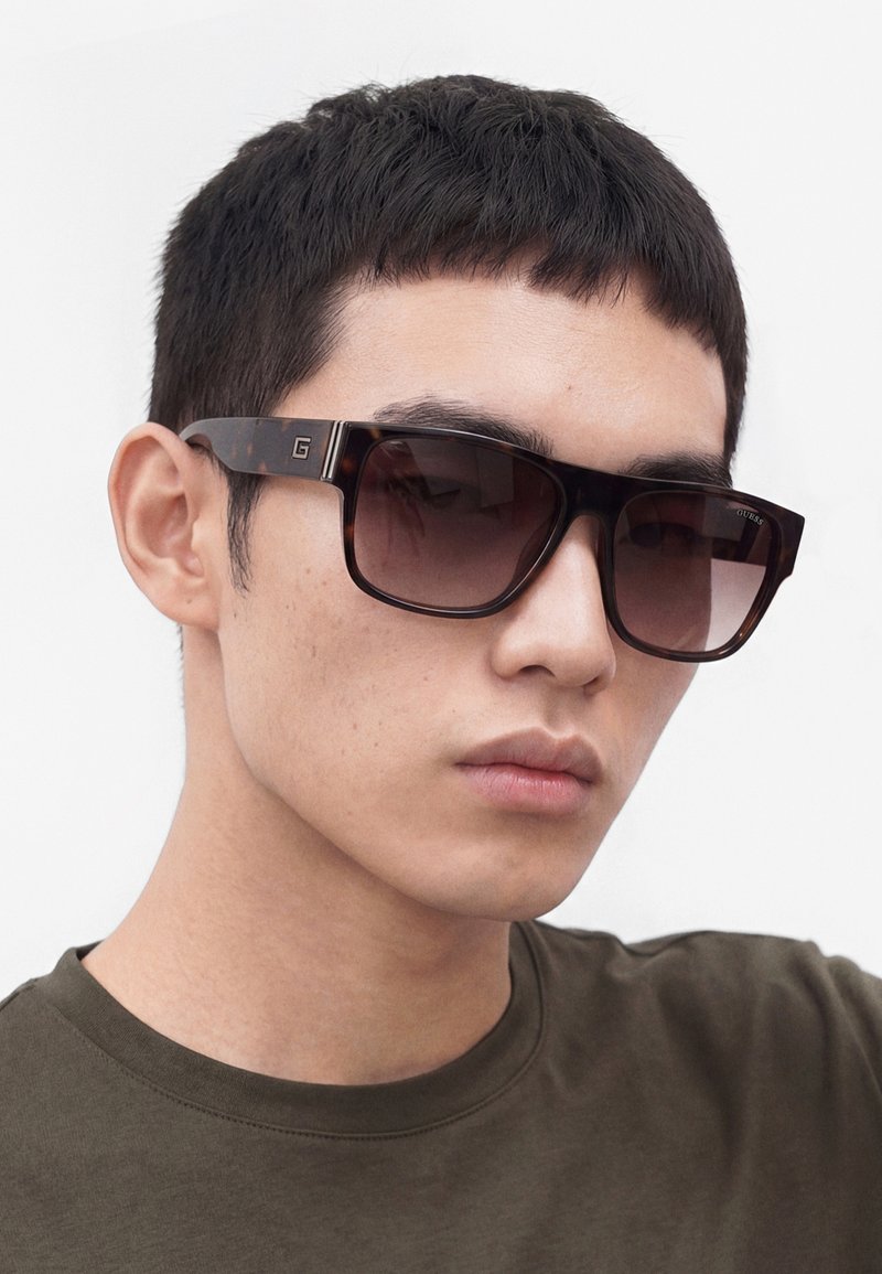 Jeune homme aux cheveux noirs courts portant des lunettes de soleil rectangulaires foncées et un t-shirt kaki à col rond sur un fond uni.
