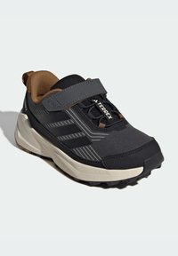 Chaussure de sport noire et grise avec une tige synthétique robuste, une sangle Velcro, des matériaux texturés, une semelle en caoutchouc légère et des accents bandés.