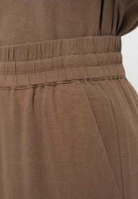 Καφέ ύφασμα με απαλή υφή· διαθέτει ελαστική μέση με πιέτες και χαλαρές πτυχές για επιπλέον ροή.