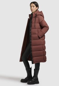 Ilgs, pūkains stils mētelis dziļi. burgundijas krāsā ar kapuci, ar rāvējslēdzēju priekšpusē un sānu kabatām; tam ir gluda tekstūra un strukturēta forma.