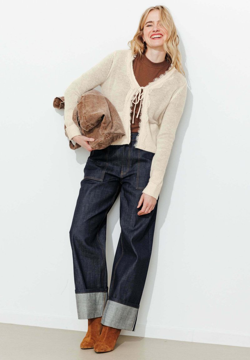 Femme souriante appuyée contre un mur, tenant une veste marron pliée, portant un cardigan crème, un haut marron, un jean foncé retroussé et des bottines camel.