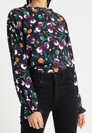 Blouse noire à fleurs avec des fleurs violettes, blanches et orange, manches longues bouffantes, poignets à volants, associée à un pantalon noir taille haute.