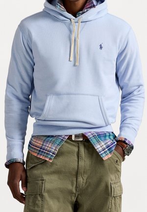 Homme portant un sweat à capuche bleu clair avec des cordons blancs, une chemise à carreaux en dessous, un pantalon cargo olive et une montre-bracelet.