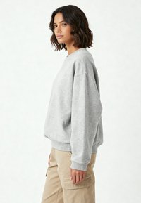 Felpa grigia chiara realizzata in tessuto morbido e testurizzato. Presenta una vestibilità rilassata, polsini a costine e un collo girocollo. Indossata con pantaloni beige.