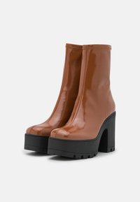 Brown patent leather ankle boots with a chunky black platform heel and a smooth, glossy finish. Features a seamless design and rounded toes.