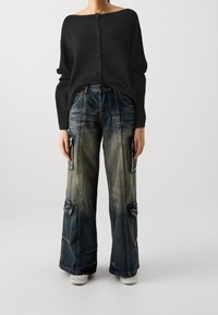 Musta ylisuurikokoinen neulepusero, jossa napit pääntiellä, yhdistettynä leveälahkeisiin revittyihin farkkuhousuihin, joissa on useita taskuja ja vaaleita haalistumia.