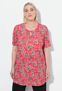 Červená tunika s paisley vzorem, krátkými rukávy a detaily v oblasti krku ve tvaru klíčové dírky. Obsahuje barevné květinové a paisley vzory po celé ploše.