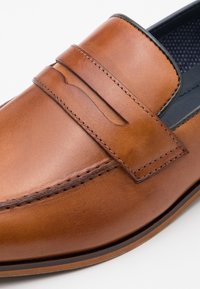 Mocassin en cuir marron avec détail de bride pour pièce et coutures visibles le long des bords et autour de la zone des orteils.
