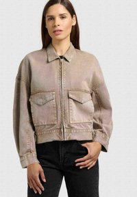Femme portant une veste courte beige à fermeture éclair avec de grandes poches devant et un pantalon noir, debout devant un fond clair uni.