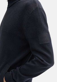 Granatowy sweter z dzianiny, z fakturowaną górną częścią, gładką dolną częścią i prostokątną metką materiałową na rękawie.