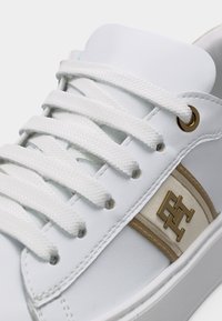 Λευκό αθλητικό παπούτσι με λευκά κορδόνια, χρυσή τρύπα για κορδόνι και χρυσλό μεταλλικό λογότυπο σε μπεζ λωρίδα στο πλάι.