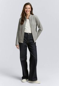 Grijze zip-up jas met een gestructureerde afwerking, gecombineerd met donkerblauwe jeans met brede pijpen en lichtgekleurde schoenen. De look is casual en eigentijds.