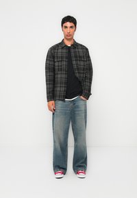 Young man standing with hands in pockets wearing black and gray plaid jacket, loose blue jeans, and red sneakers against white background.