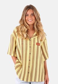 Chemise à manches courtes rayée jaune et marron avec un devant à boutons, présentant un petit logo brodé et un matériau texturé et respirant.