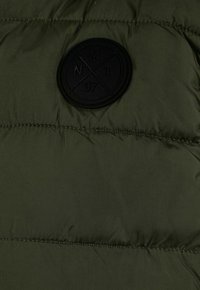 Doudoune vert olive, présentant un tissu texturé, des panneaux cousus et un patch circulaire noir avec des détails embossés.