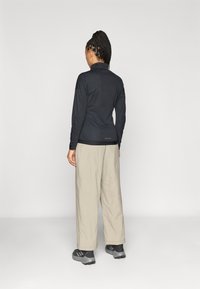 Μαύρη μπλούζα με μακριά μανίκια και κλείσιμο με φερμουάρ στον γιακά, σε συνδυασμό με ανοιχτό μπεζ παντελόνι και γκρι παπούτσια. Διαθέτει εφαρμοστή γραμμή και υφή υφάσματος.