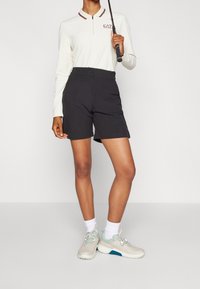 Osoba oblečená v bílém polo tričku s dlouhým rukávem, černých šortkách, bílých ponožkách a béžových teniskách, držící golfovou hůl přes rameno.