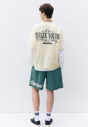 Jeune homme portant une chemise beige avec le texte « ESTATIC YOUTH but Always Evolving », un short vert à carreaux avec le texte « Slow », des chaussettes blanches et des chaussures noires, de dos.