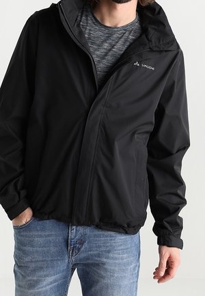 Mann trägt schwarze Vaude-Kapuzensjacke über grauem Hemd und blauer Jeans, steht mit einer Hand in der Tasche vor weißem Hintergrund.