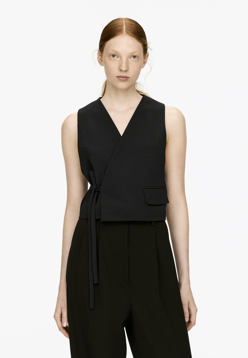 Veste noire à enrouler avec décolleté en V profond, liens sur les côtés et une poche avant, associée à un pantalon sombre taille haute, doté d'une texture de tissu lisse.
