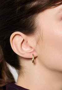 Złoty kolczyk w kształcie obręczy pokazany na uchu, posiadający gładkie, wypolerowane wykończenie oraz okrągły kształt, zawieszony na małym zapięciu.