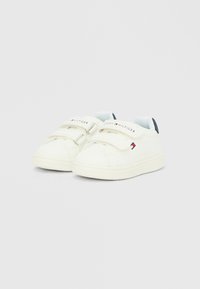 Λευκά αθλητικά παπούτσια για νήπια με δύο λουριά βέλκρο, διακριτικό λογότυπο Tommy Hilfiger στα λουριά και πλάι, και μπλε ταμπελίτσα στο τακούνι σε μονόχρωμο φόντο.