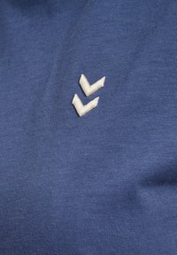 Deux chevrons blancs brodés sur un fond en tissu bleu texturé, centrés dans l'image.