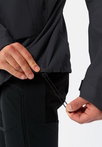 Giacca nera leggera con orlo ed elastici ai polsini; mani che regolano il cordino in vita; superficie liscia, design minimalista.