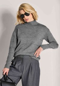 Frau mit blonden Haaren, die schwarze Sonnenbrille, einen grauen Rollkragenpullover, graue Nadelstreifenhosen trägt und eine schwarze Handtasche hält.