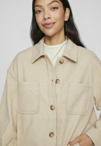 Giacca in misto lana beige con colletto, chiusura frontale con bottoni e due grandi tasche applicate. Texture morbida con un motivo sottile.