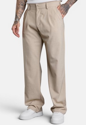 Mann trägt beige weite Hose, weiße Sneaker und ein weißes Hemd mit sichtbaren tätowierten Armen, eine Hand in der Tasche.