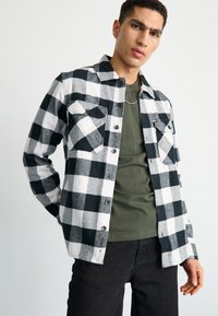 Camisa de franela a cuadros en blanco y negro con dos bolsillos frontales, botones negros, superpuesta a una camiseta verde oscuro, combinada con pantalones negros.