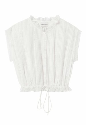 Femme portant un chemisier blanc sans manches, un pantalon beige à jambes larges avec une ceinture noire, des chaussures beiges et tenant un sac à main beige.