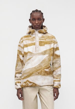 Personne portant un pull en polaire beige et blanc à motifs avec capuche et boutons-pression, associé à un pantalon beige, debout devant un fond uni.