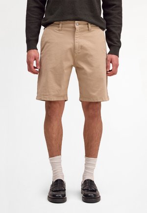 Mężczyzna w beżowych szortach do kolan, białych skarpetach do kostek, czarnych skórzanych mokasynach i ciemnym, prążkowanym swetrze z długim rękawem stoi na jednolitym tle.