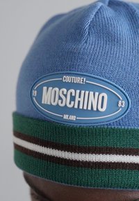 Caciula tricotată albastră cu o margine dungată verde și alb. Prezintă o patch circulară albastră cu "MOSCHINO" și "COUTURE! MILANO" în litere îngroșate.