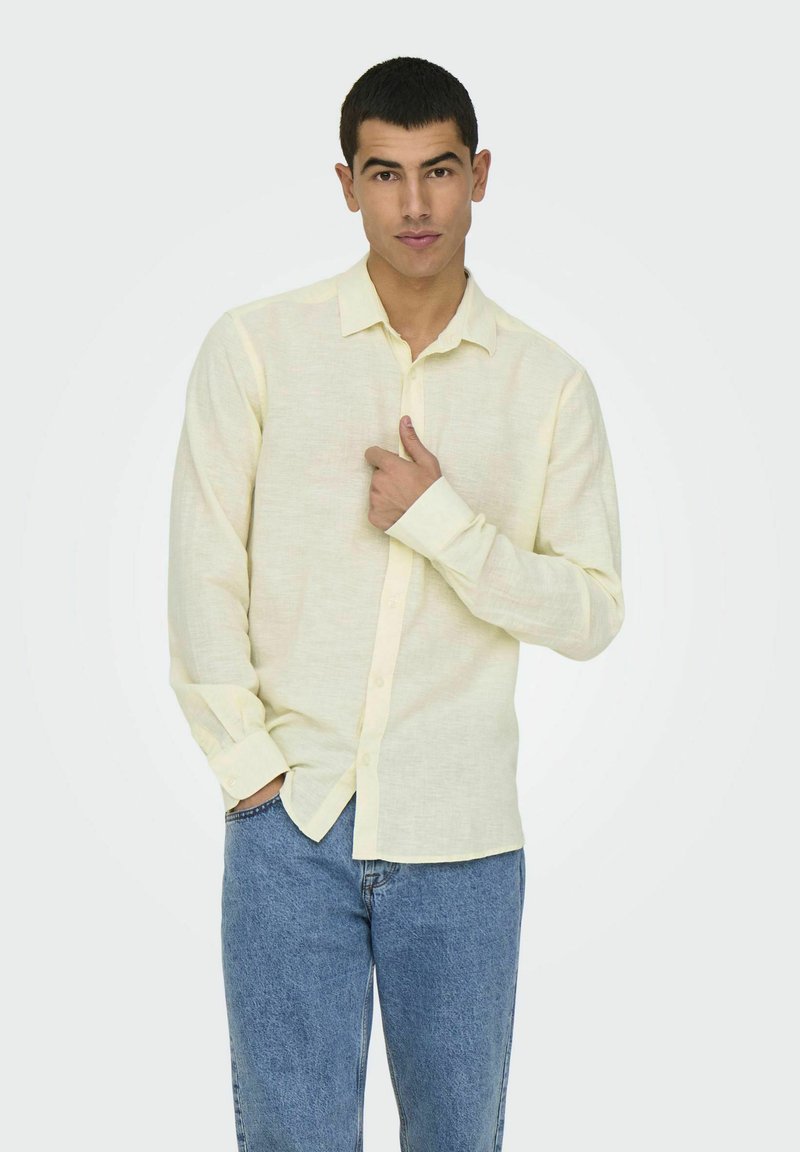 Chemise longue à manches jaunes claires avec un col boutonné, faite de tissu texturé, associée à un jean bleu. Le modèle se tient avec la main sur la poitrine.