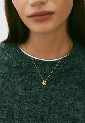 Zelta kaklarota ar mezgla piekariņu, valkāta virs teksturēta tumši zaļa džempera ar ribotu apkakli un baltu apakškārtu.