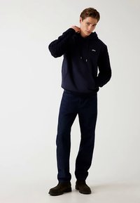 Marineblauer Kapuzenpullover mit strukturierten, schwarzen Rippärmeln und Kordeln, kombiniert mit dunkelblauen Jeans und braunen Schuhen. Einfaches Design, lässige Passform.