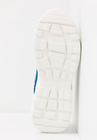 Semelle de sneaker en caoutchouc blanc avec un motif texturé pour l'adhérence, avec une partie supérieure en textile bleu et gris fixée de manière visible sur les côtés.