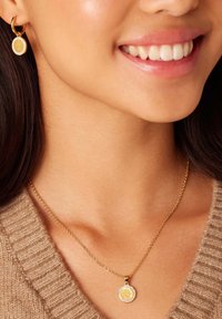 Gouden oorbellen en een bijpassende ketting hebben ronde hangers met een handsymbool, versierd met rhinestones, tegen een gebreide bruine trui.