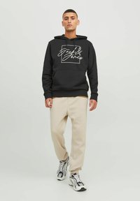 Schwarzer Hoodie mit weißem Logo, vorderer Beuteltasche und Kapuze mit Kordelzug. Kombiniert mit beigen Jogginghosen und grauen Sneakers. Schlicher Hintergrund.