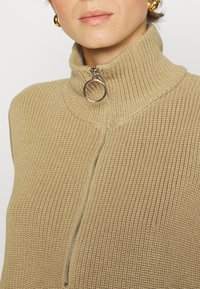 Beige korkeakauluksinen neule, jossa on teksturoitu neule, puolivetoketju edessä ja ympyräinen hopeinen vetoketjun pää, sekä ribbireunukset kauluksessa.