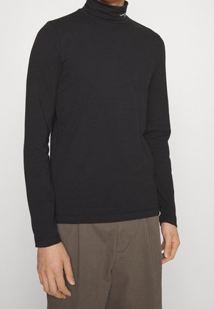Personne portant un col roulé noir à manches longues et un pantalon marron, debout devant un fond clair uni.